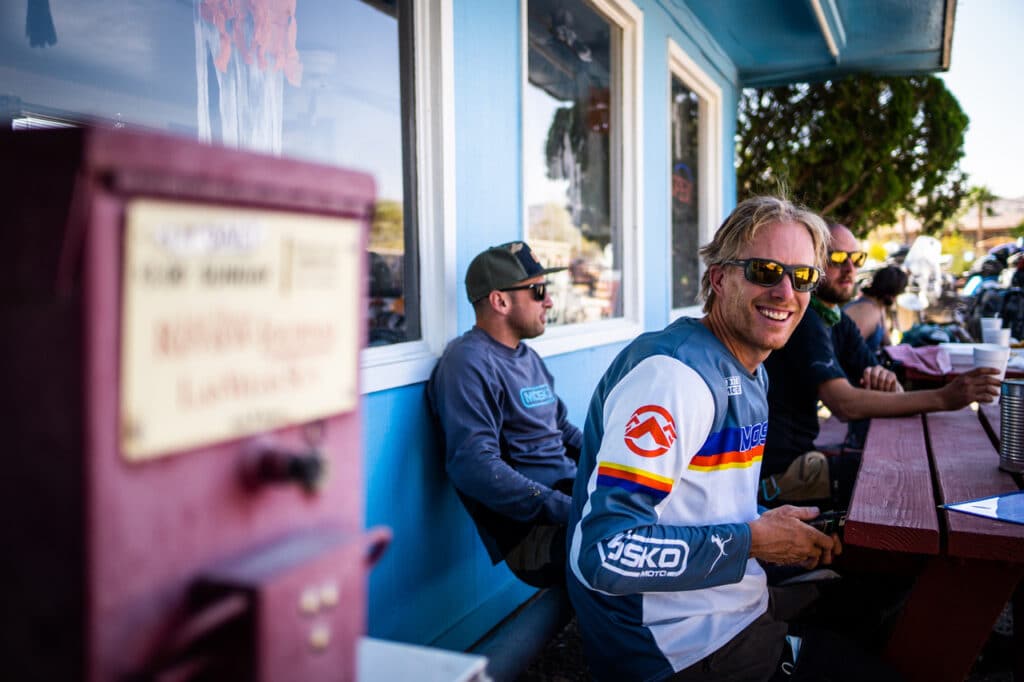 People sit together at a wooden picnic table outside a blue building, sharing a relaxed moment. One person smiles toward the camera while holding a drink, with others nearby, sunlight reflecting off windows and motorcycles gathered in the background.
