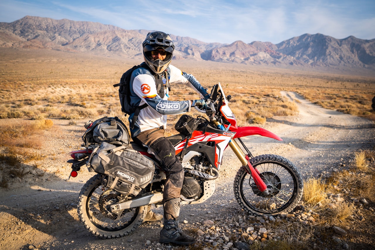 A person sits on a loaded off-road motorcycle on a gravel trail, wearing a helmet and riding gear. Desert plains stretch outward toward layered mountain ridges beneath a wide, open sky.