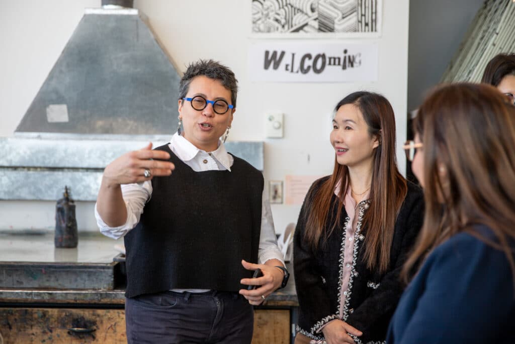 A person gestures while speaking to a small group in a studio workspace with equipment and artwork in the background.