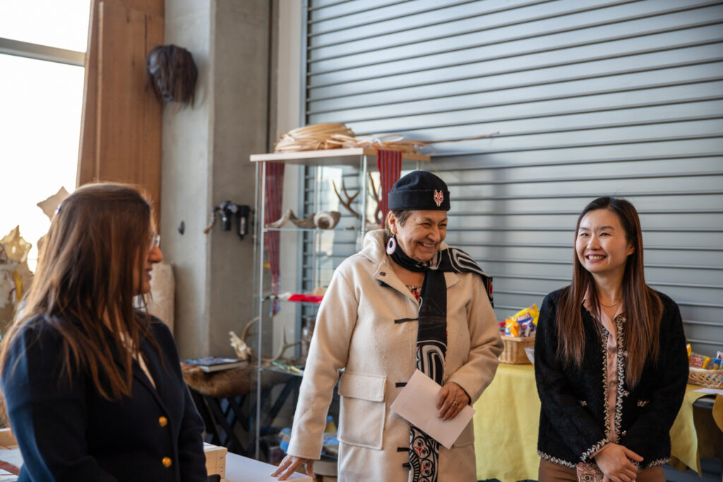 Three people stand together smiling and talking in a workspace with display shelves and materials nearby.