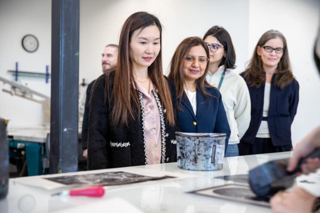 Several people stand around a worktable watching a demonstration involving printed materials and tools in a workshop setting.