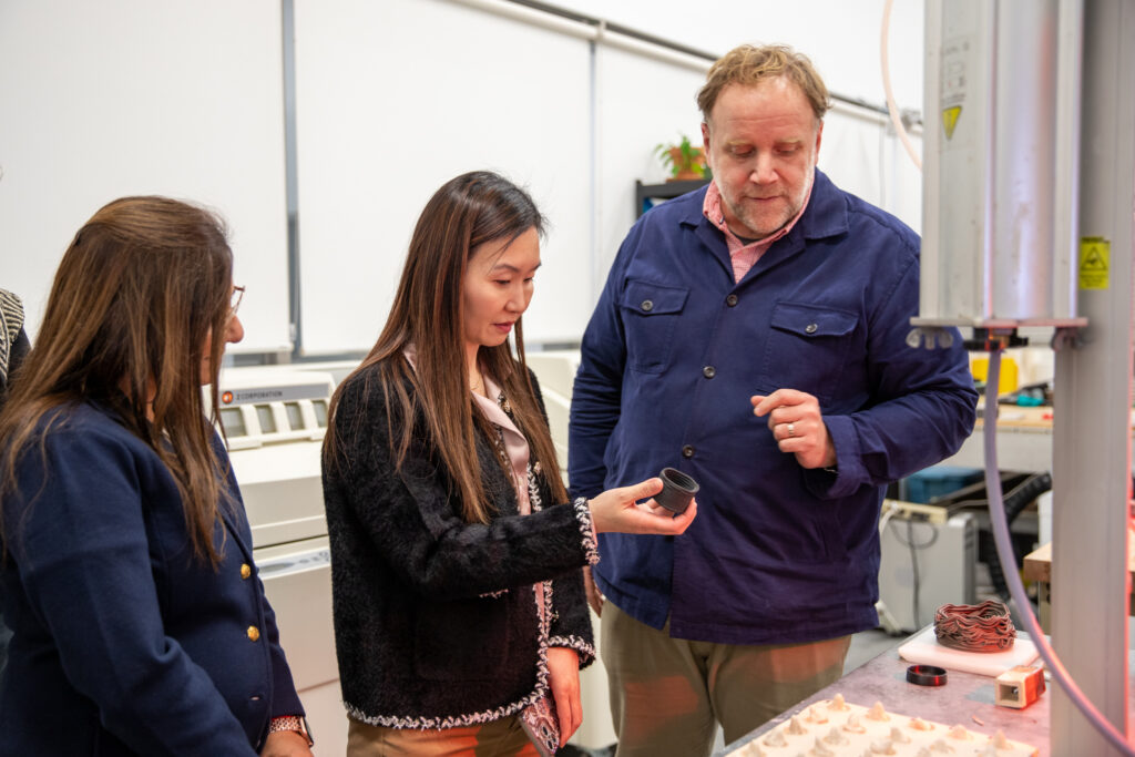 Three people examine a small object beside industrial equipment in a fabrication lab.