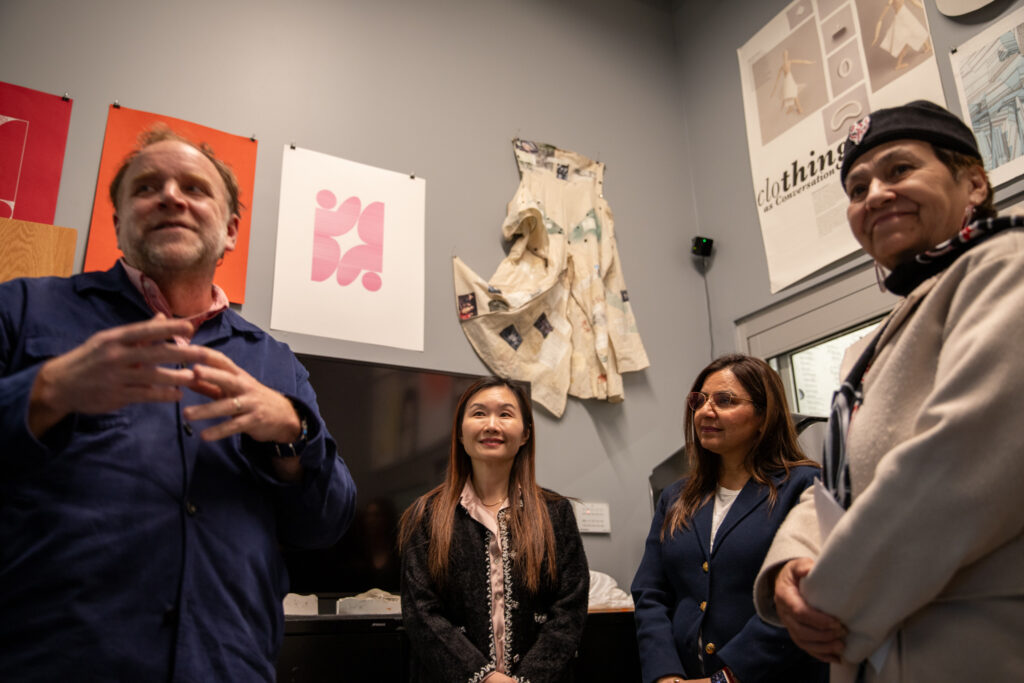 A small group listens to a speaker in a studio where garments and design posters hang on the walls.