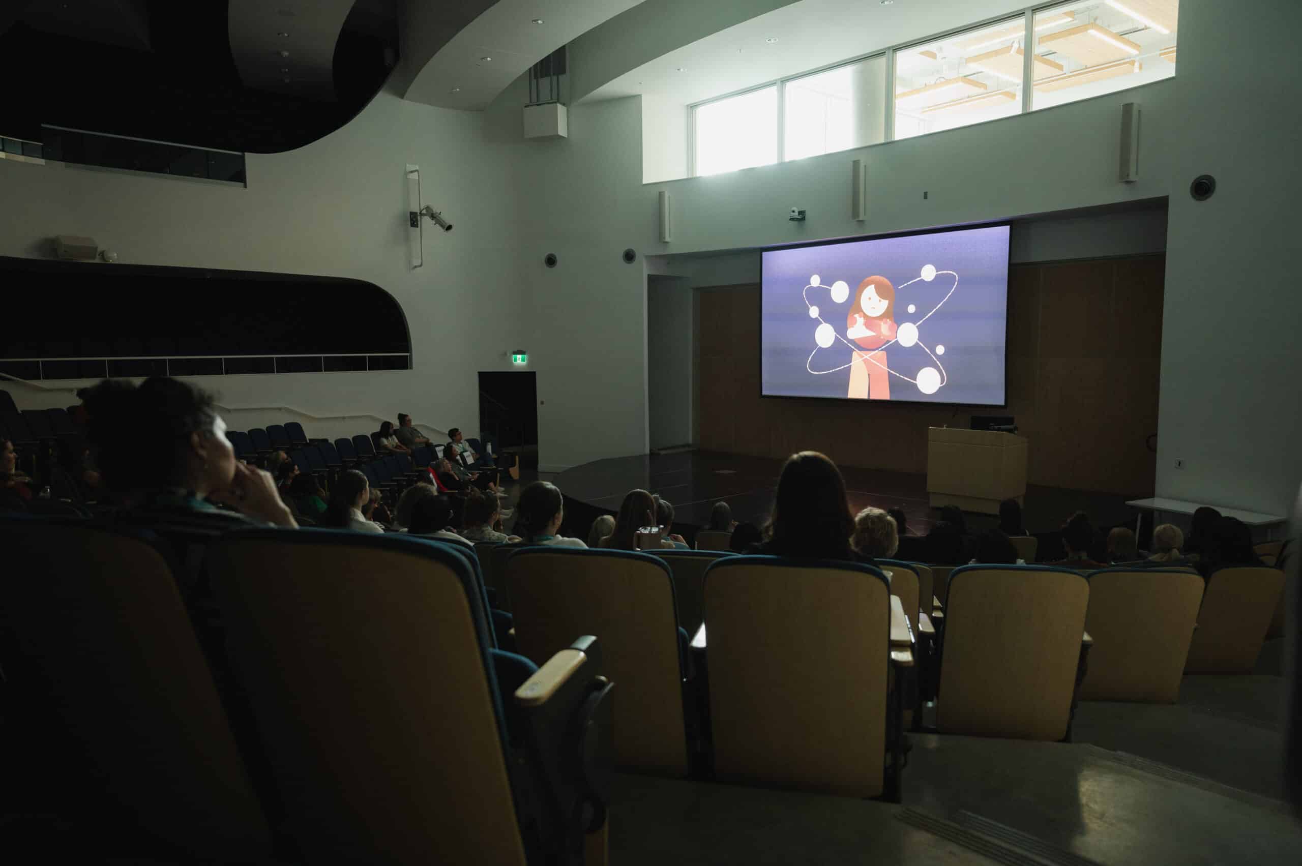An audience sits in a softly lit auditorium, facing a large screen where an animated figure appears, creating a quiet, collective focus as the room gathers around a shared story.