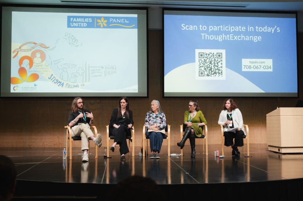 A panel of five people sits on a stage in a lecture hall, mid-conversation, with projected slides behind them inviting audience participation, capturing a shared moment of listening and dialogue.