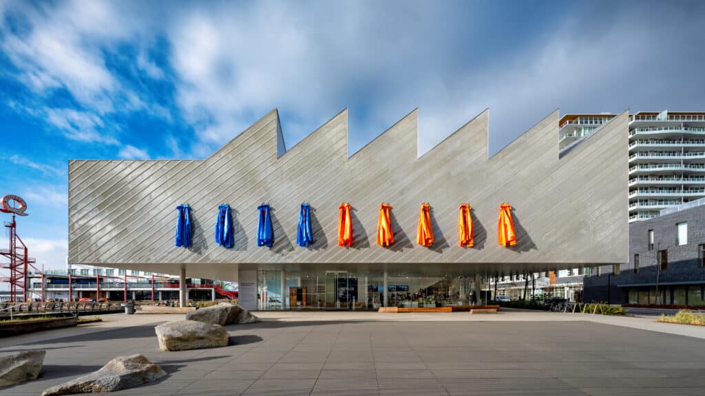 A modern gallery building stands beneath an open sky, its angular facade lined with blue and orange hanging garments arranged in a steady rhythm.