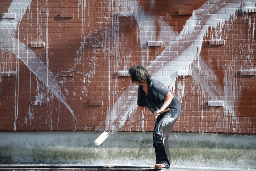 A person swings a bucket of paint against a brick wall, sending a splash through the air in a moment of motion.