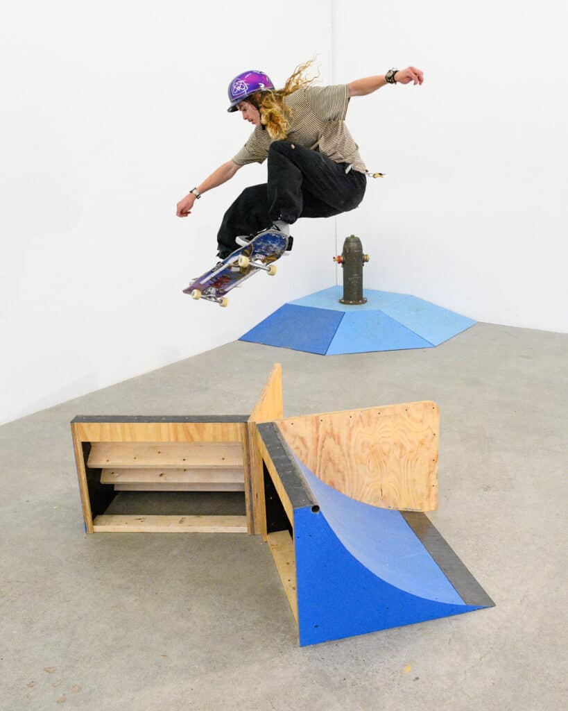 A person jumps a skateboard off a ramp into the air inside a bright studio, clearing a small wooden ramp. Blue and plywood surfaces shape the space below, with a simple sculptural form set against white walls.