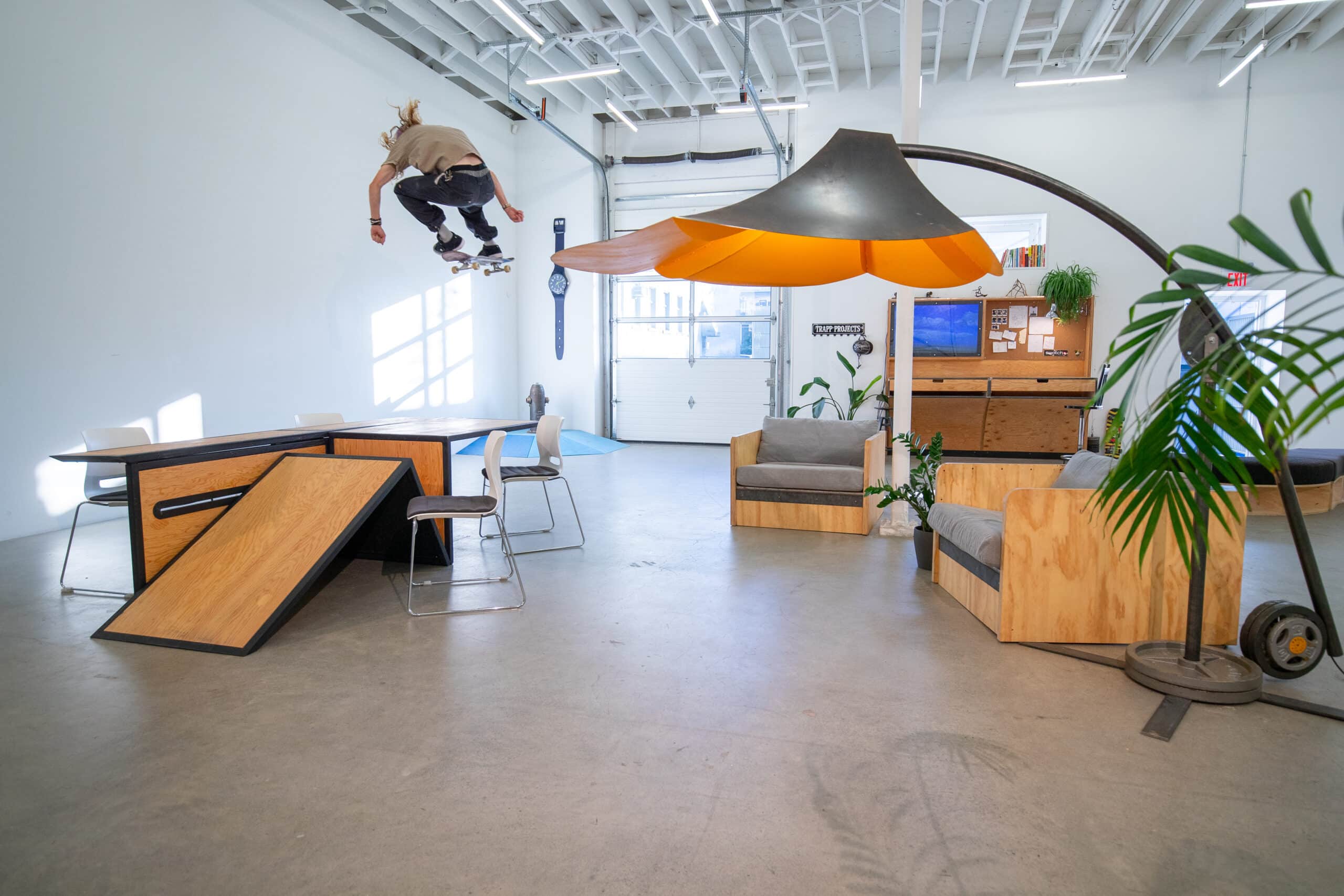 A person launches a skateboard into the air inside a bright studio space. Ramps, tables and seating form an improvised landscape below, while light pours in from high windows and an oversized lamp curves across the room.