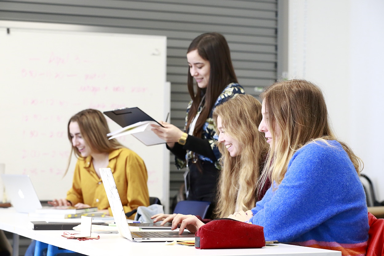 A small group of students work side by side at a table, smiling and focused on laptops and notes in a bright classroom setting.