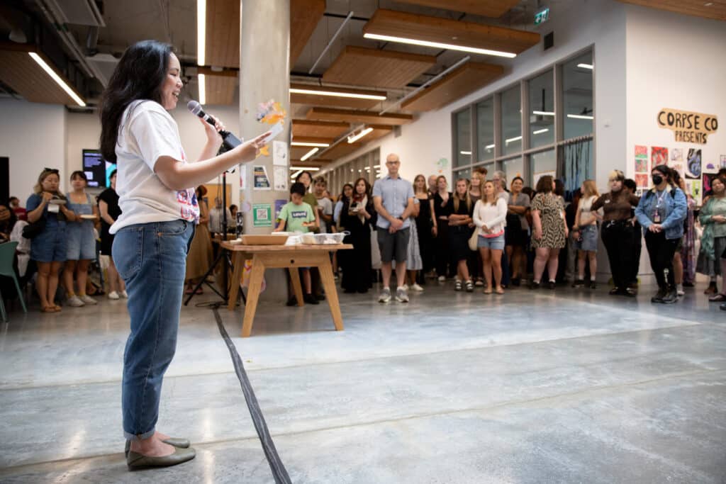 A person speaks into a microphone in a spacious, brightly lit gallery space surrounded by a crowd of people.