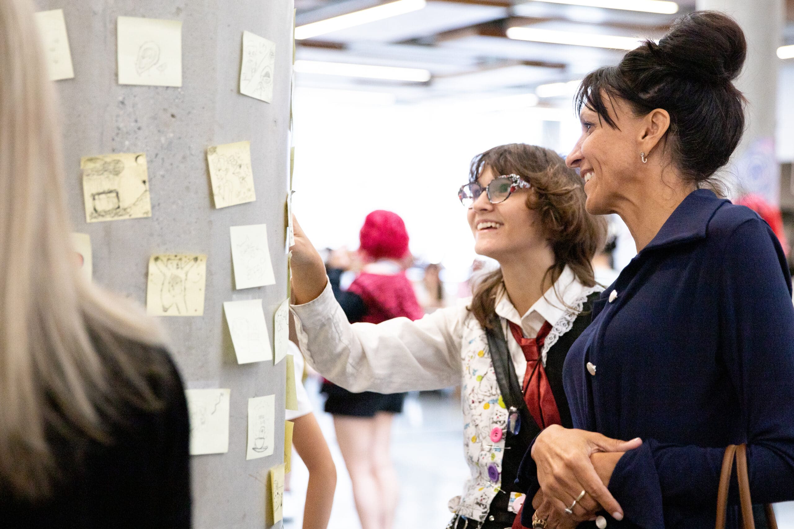 A young person and an adult stand shoulder to shoulder smiling and pointing as they view artworks adhered to a concrete post in a brightly lit gallery.