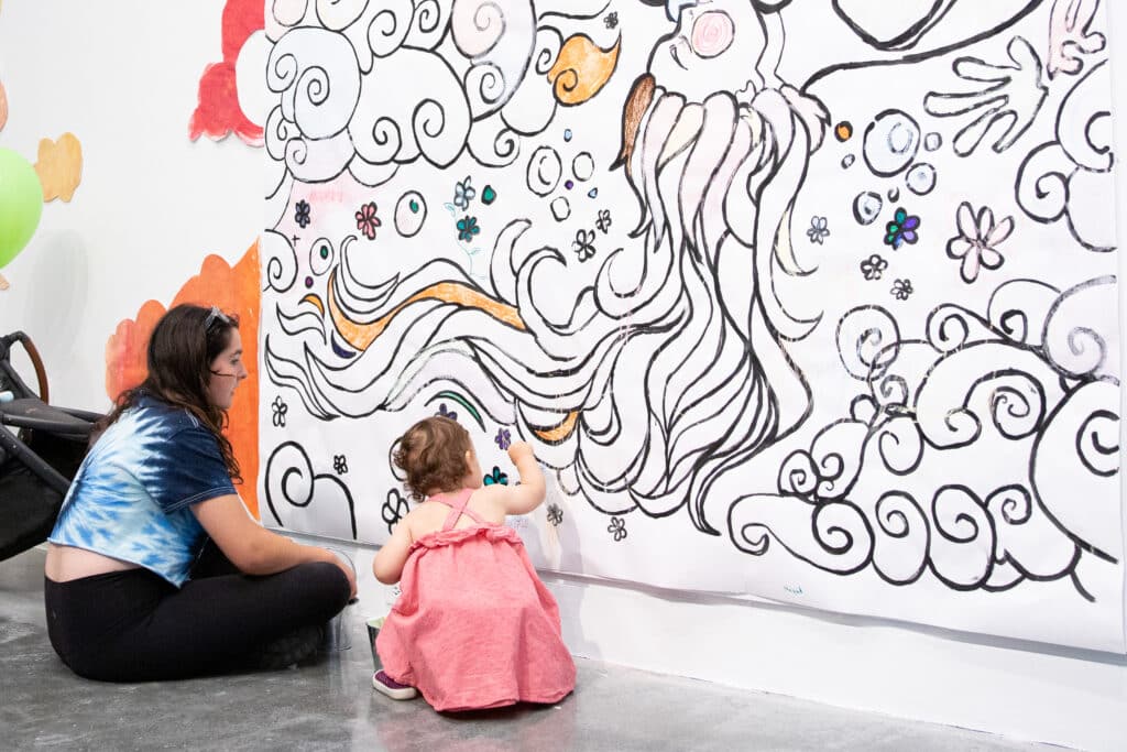 A parent and a young child sit on the concrete floor of a gallery exploring a large drawing with swirling lines and pops of bright colour.