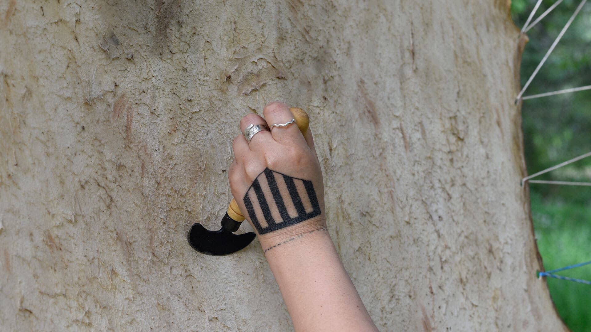 A hand presses a curved tool into a textured hide surface, carving or shaping the material. Rings and a geometric tattoo are visible as the tool moves slowly across the natural grain.