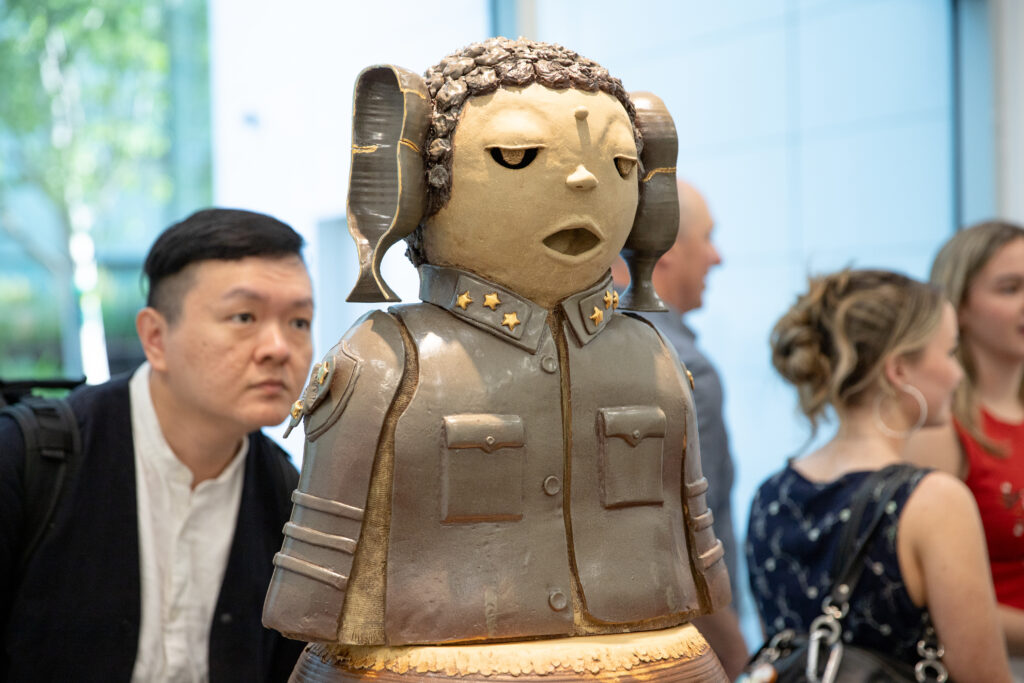 A ceramic sculpture of a stylized child in a military-style jacket is displayed in a gallery space as visitors look on in the background.