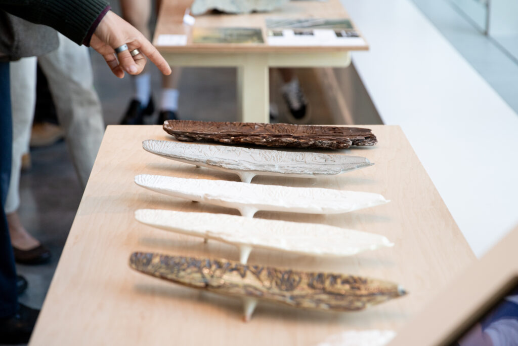 Handmade ceramic forms in white and earthy tones are arranged in a row on a wooden display table as someone points toward them.