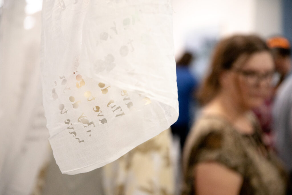 A translucent textile artwork with faint printed text hangs in the foreground while visitors move through the gallery behind it.