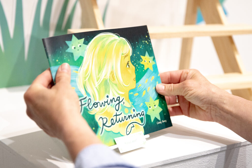 Hands hold an illustrated book titled Flowing, Returning, featuring a profile portrait of a child against a star-filled green background.