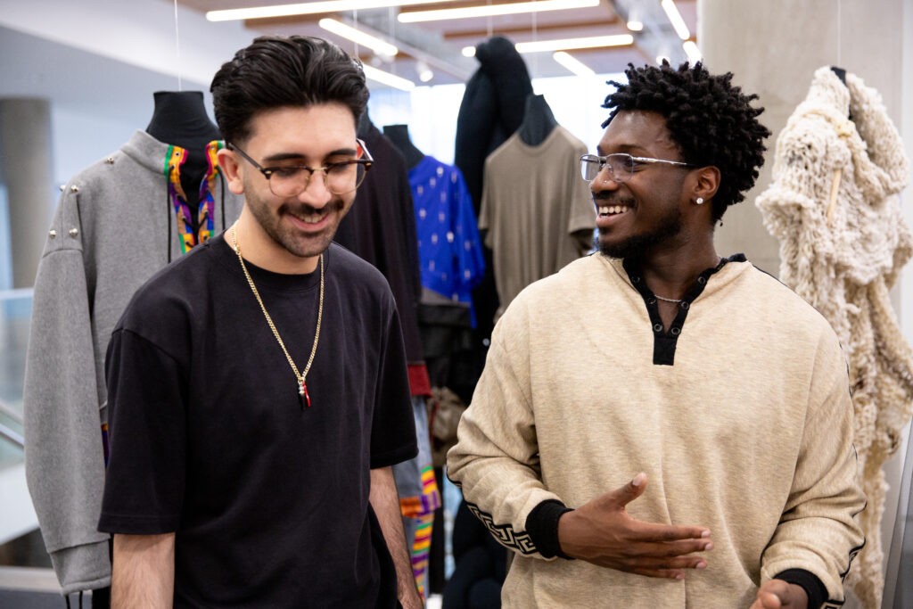 Two people smile and talk in a fashion display area, standing among mannequins dressed in contemporary garments and textured knitwear.