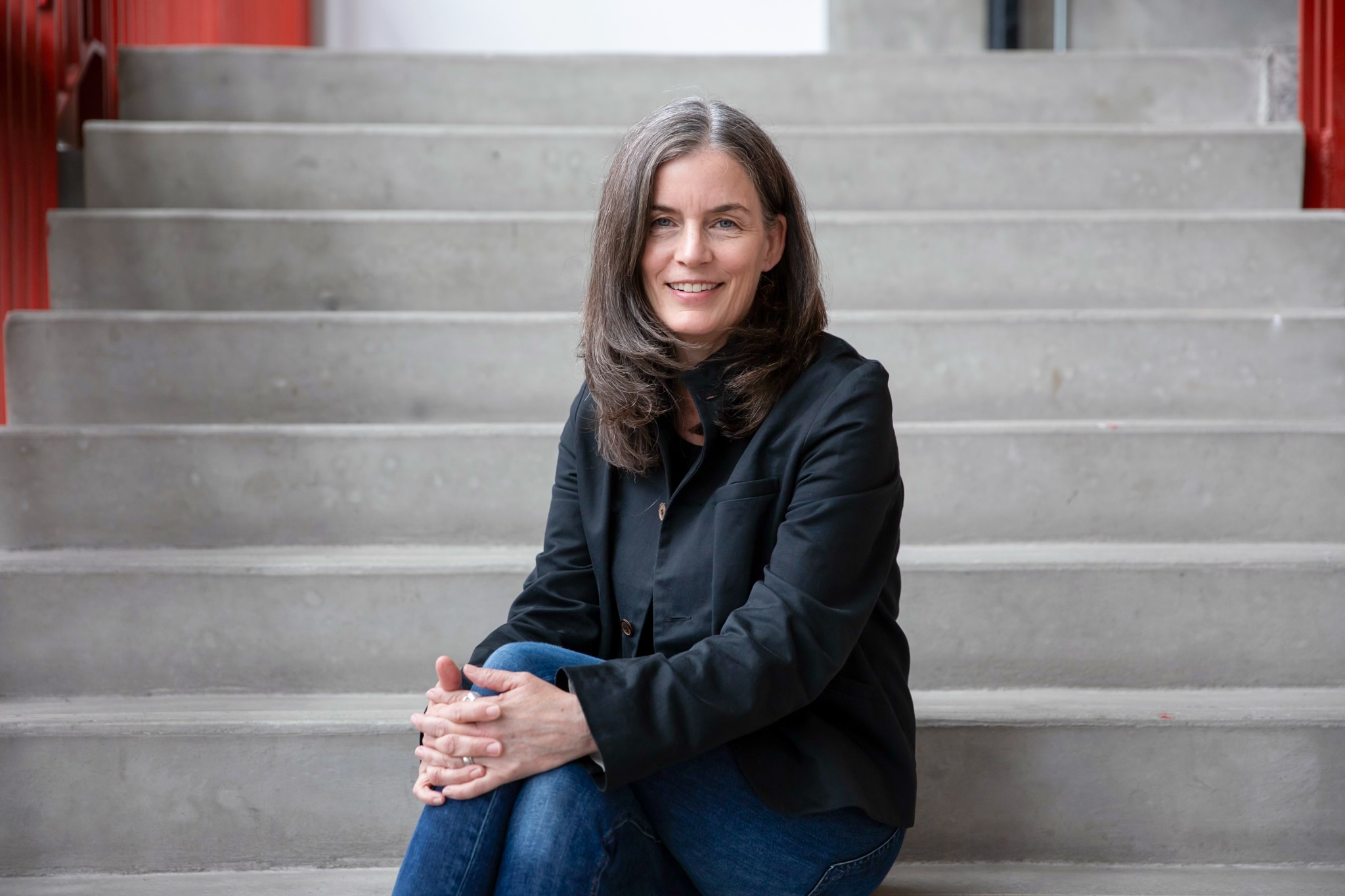A person sits on wide concrete steps, hands loosely clasped, wearing a dark jacket and jeans, offering a relaxed smile as soft light settles across the stairway behind them.