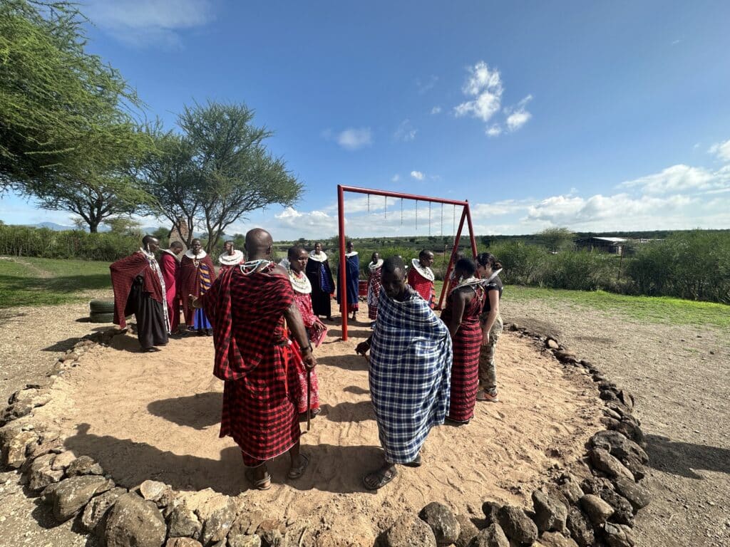 A group of people stand in a circle on a sandy clearing, wearing patterned garments in red, blue and black. A swing frame rises at the centre, with trees, open land and a wide blue sky stretching into the distance.