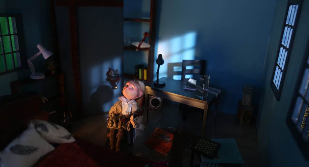 A small figurine sits holding a violin in a dimly lit room. Blue light filters through a window onto a desk, lamp and shelves, creating a quiet, staged interior with books and objects scattered nearby.