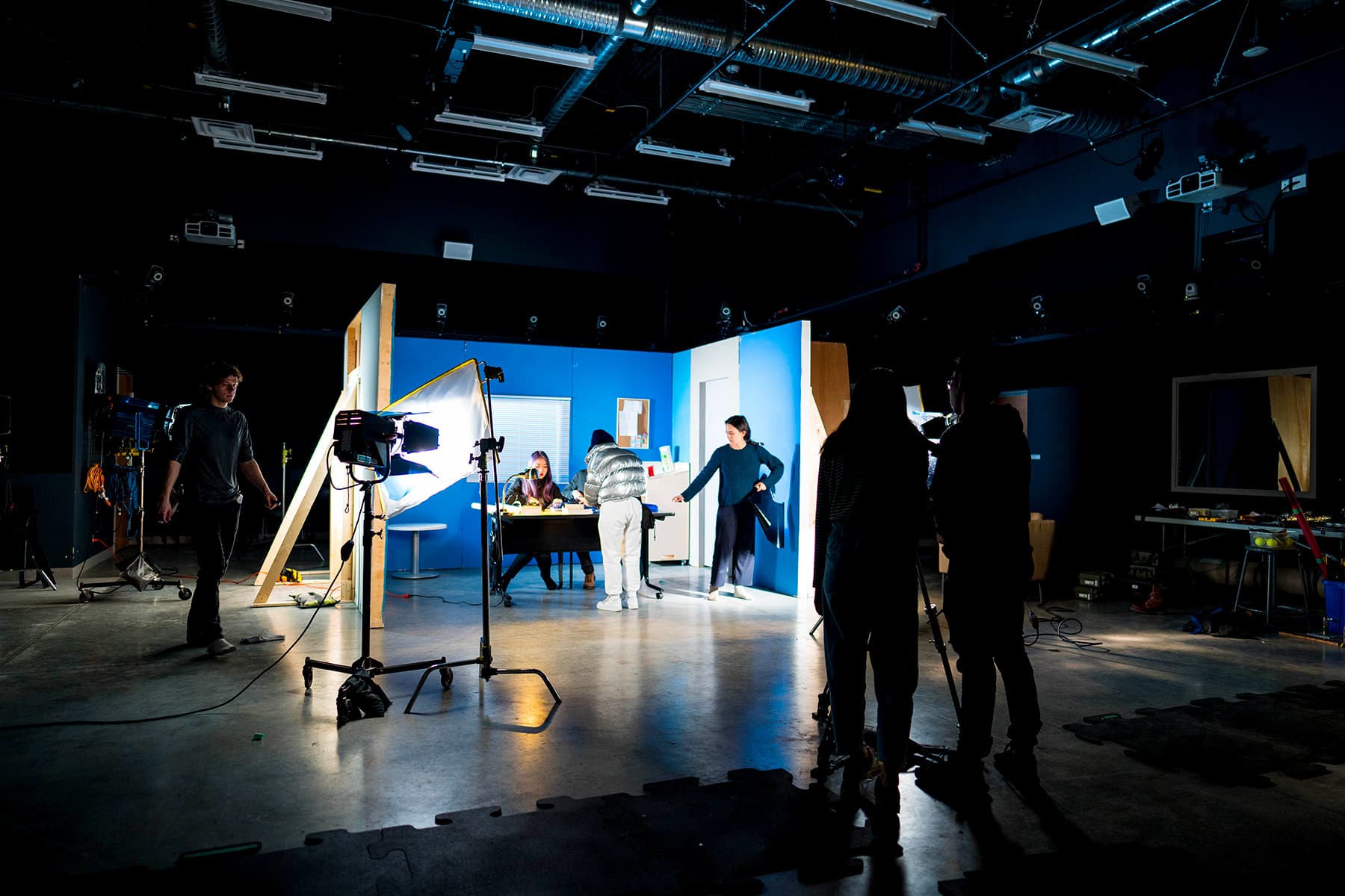 In a professional studio with exposed ceiling ducts and industrial lighting rigs, students collaborate around a blue-walled set. One individual adjusts a large softbox light, while others gather near a table in the set, with equipment like cameras, cables, and a window with blinds visible throughout the space. The scene blends shadows and artificial illumination, highlighting hands-on production work.