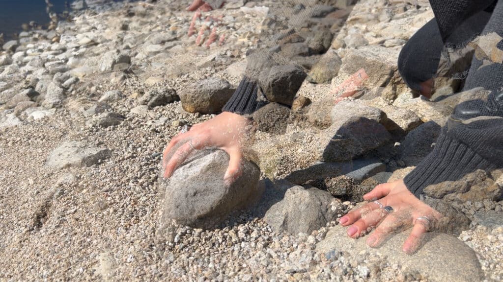 Hands press into sand and smooth stones at the water’s edge, layered with faint overlapping movement. The textures of rock, skin and shoreline blur together in soft light.