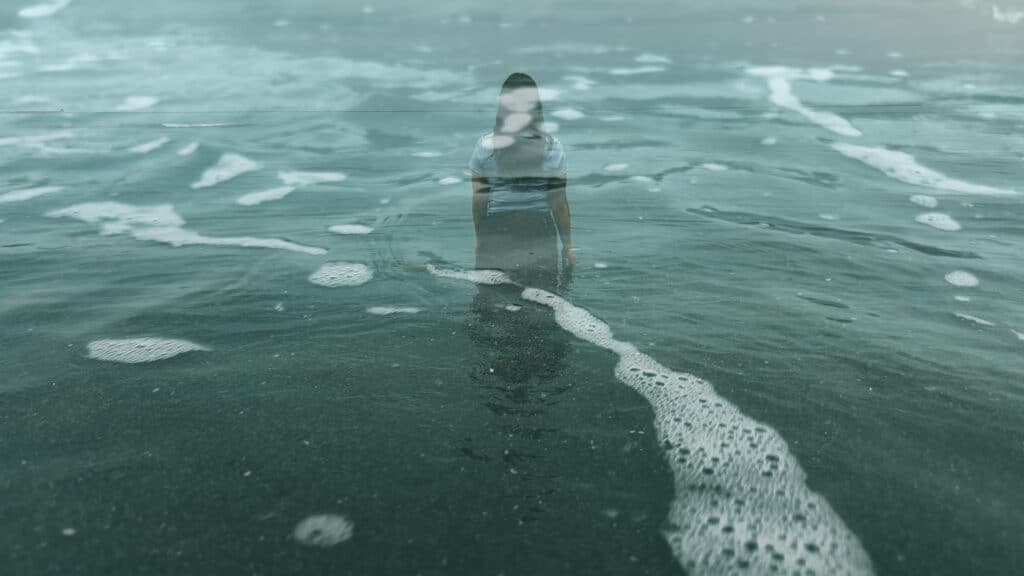 A person is superimposed across the face of open water with layered reflections of waves and sky passing across their body. The image blends figure and sea into a single, fluid moment.