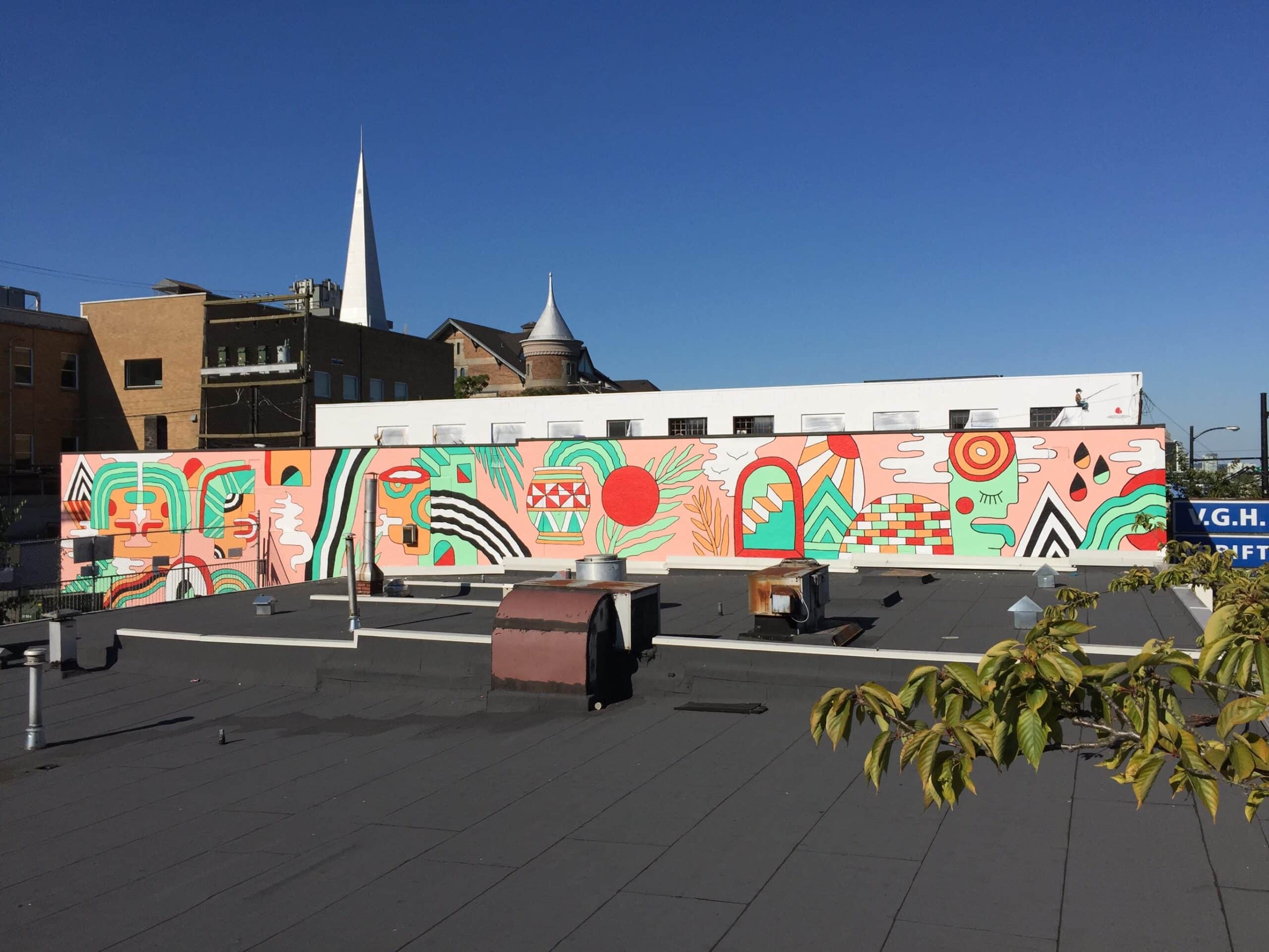 Wide rooftop view of a long, colourful abstract mural featuring geometric shapes, faces and plant forms, painted across a building wall under a clear blue sky.