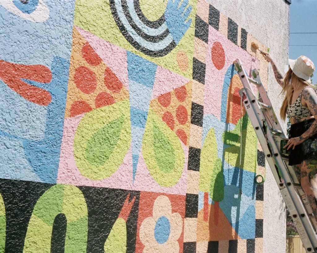 Close-up of an artist painting a geometric mural from a ladder, applying colour to a wall filled with abstract shapes, dots and organic forms.