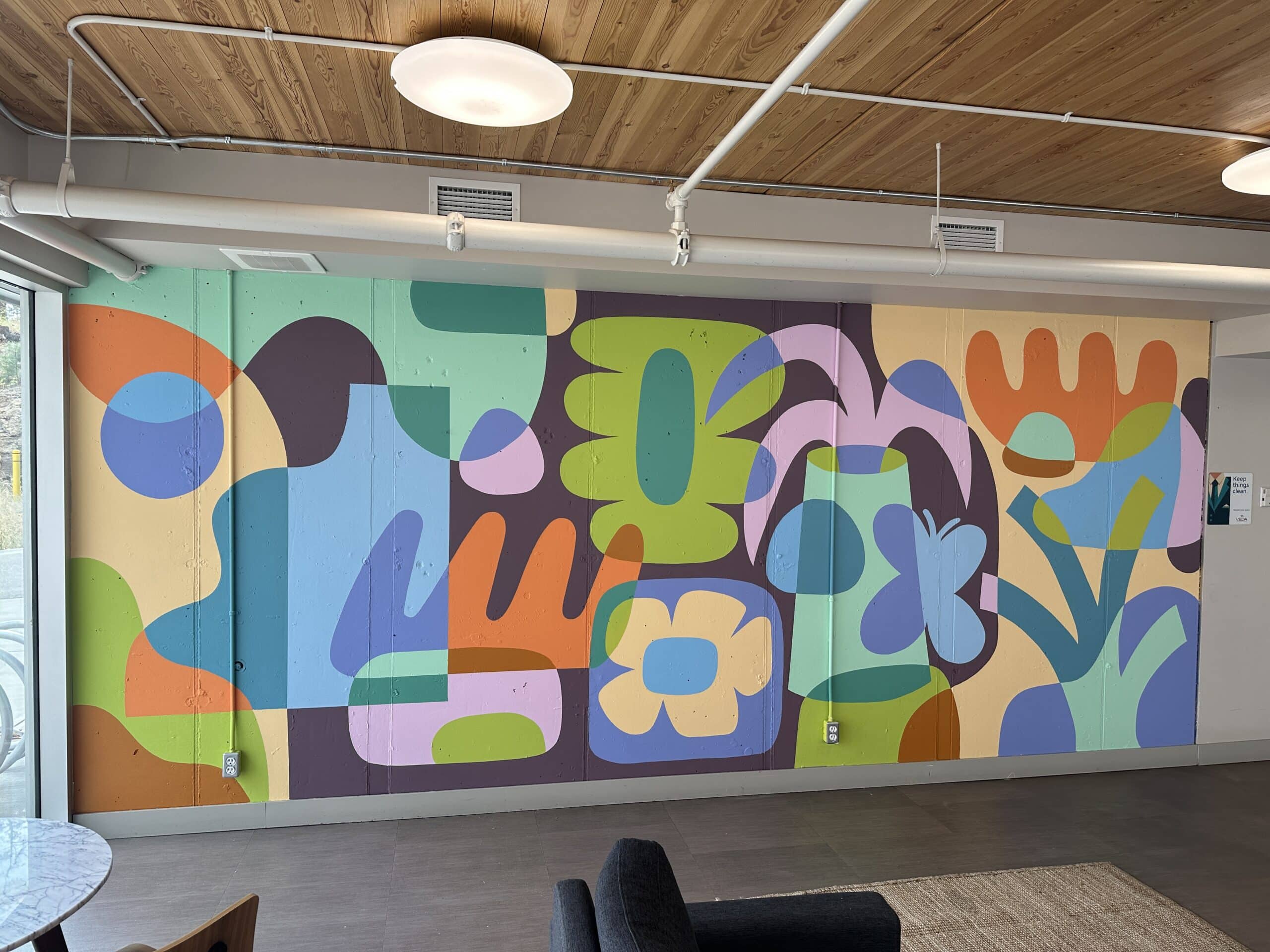 Interior wall mural spanning a lounge space, featuring large abstract plant and floral shapes in muted greens, blues, oranges and purples beneath a wood ceiling.