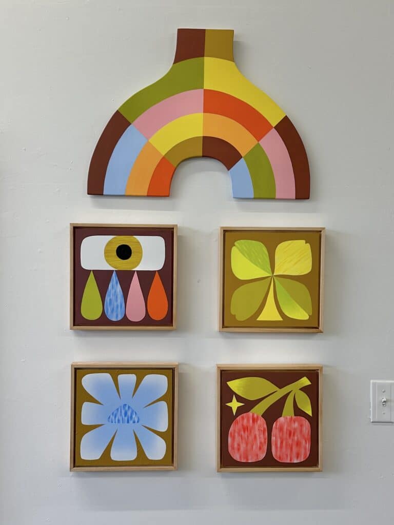 Gallery wall displaying a small sculptural rainbow above four framed graphic artworks featuring stylized eyes, flowers and fruit in warm, saturated colours.