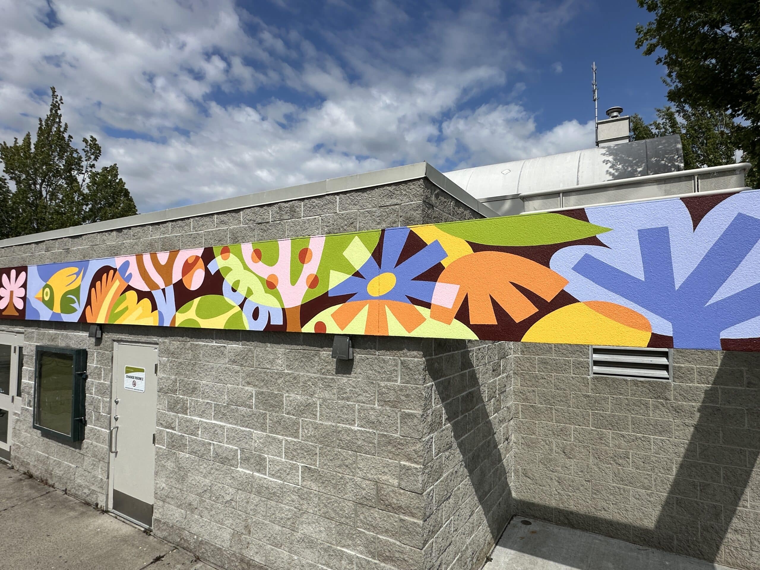 Long exterior mural wrapping around a concrete building, showing flowing abstract plant and landscape shapes in greens, blues, yellows and oranges.