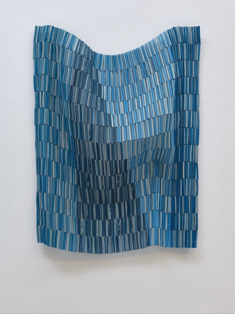 A sculptural wall work made of narrow blue strips arranged in gentle waves, forming a textured surface that ripples like fabric or water against a white gallery wall.