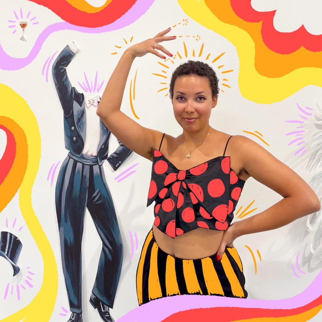 A person poses with one arm raised in a gallery space, wearing a patterned top and skirt, surrounded by colourful graphic shapes layered over a wall installation featuring costume-like cutout figures.