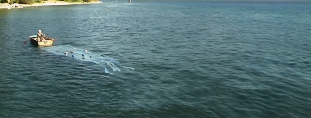 A small rowboat floats on open water, trailing a translucent sheet or net just beneath the surface, creating a faint geometric shape in the waves.