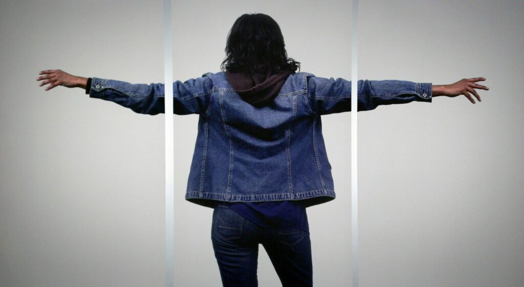 A person stands facing a plain wall with arms extended wide, their body forming a quiet cross shape within the frame.