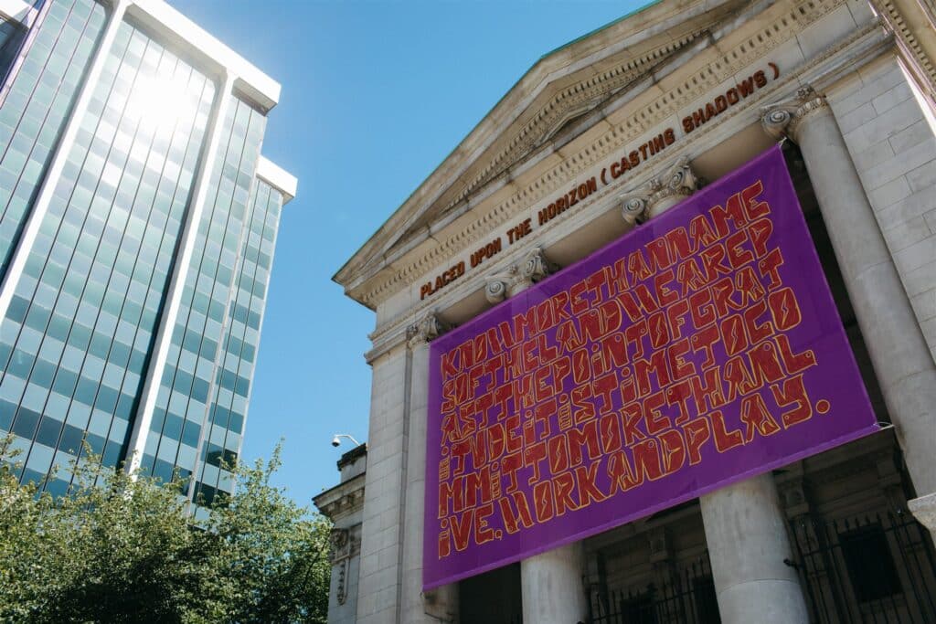 A historic stone building rises against a bright blue sky, its columns draped with a large purple banner filled with bold, swirling text. Beside it, a modern glass tower reflects the sunlight, contrasting old and new architecture.