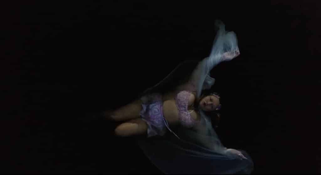 A figure floats in dark water, wrapped in sheer fabric that drifts around them. Limbs move slowly through shadow, creating a soft, suspended moment between stillness and motion.