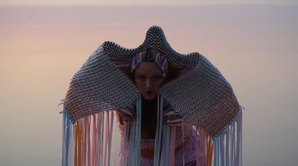 A person leans forward beneath a sculptural, woven garment that rises like a soft shell around their shoulders. Strands hang down in gentle lines as warm light fades into a quiet, dreamlike horizon.