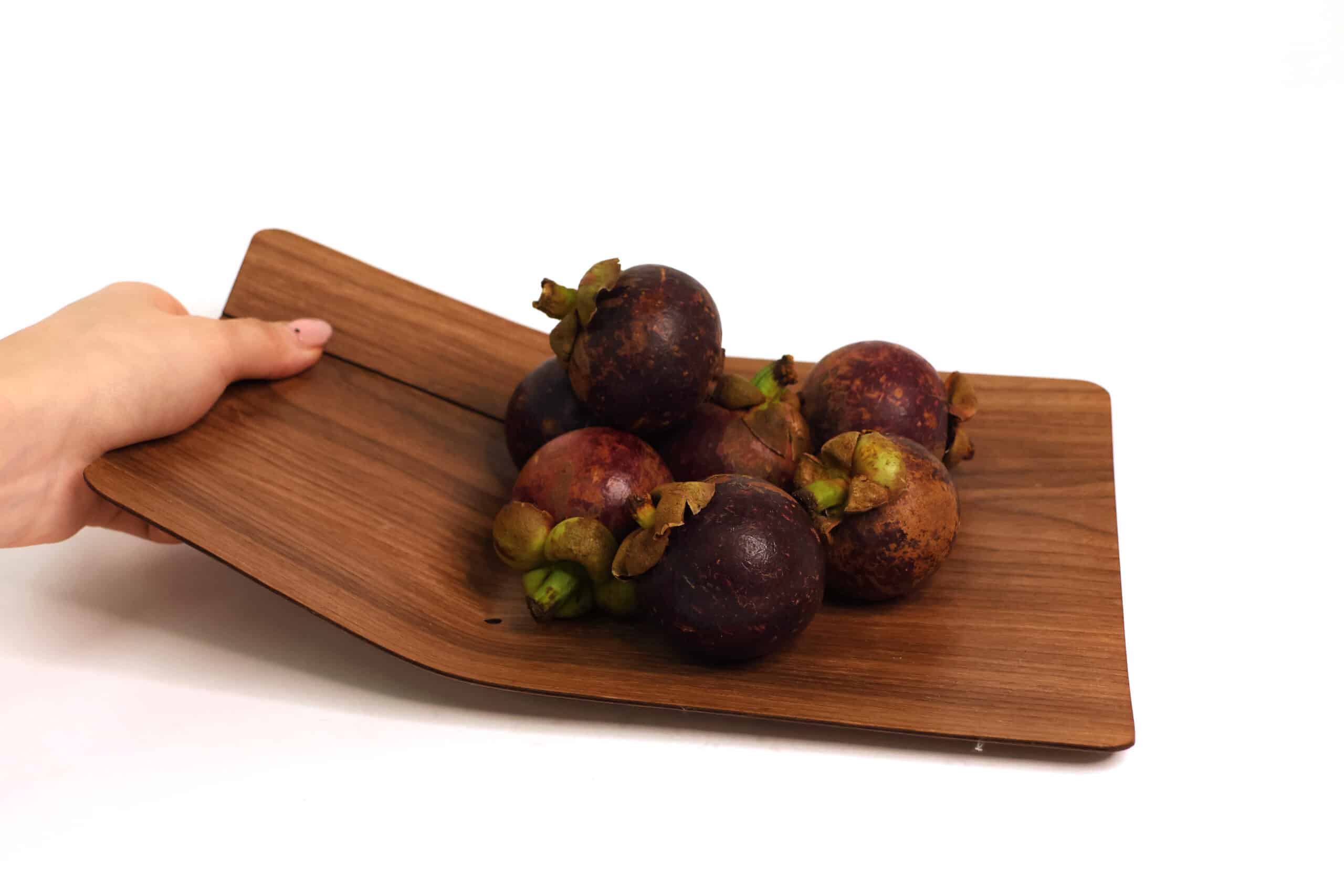 A hand tilts a curved wooden tray, gently presenting a small pile of dark purple mangosteens. The smooth grain of the wood contrasts with the fruit’s textured skins.