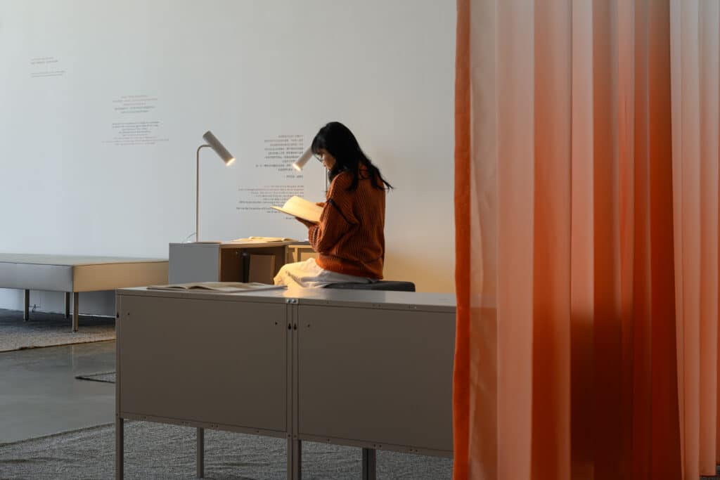 A person sits on a low bench, reading beneath a small lamp. Warm light gathers on the open pages, while a soft curtain glows nearby, creating a quiet corner that feels held in stillness.