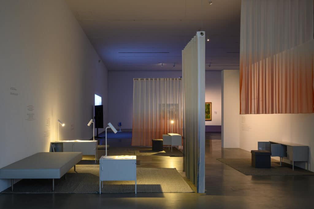 A wide view shows softly lit reading stations arranged among translucent curtains. Pools of warm light rest on books and benches, inviting slow movement and quiet attention.