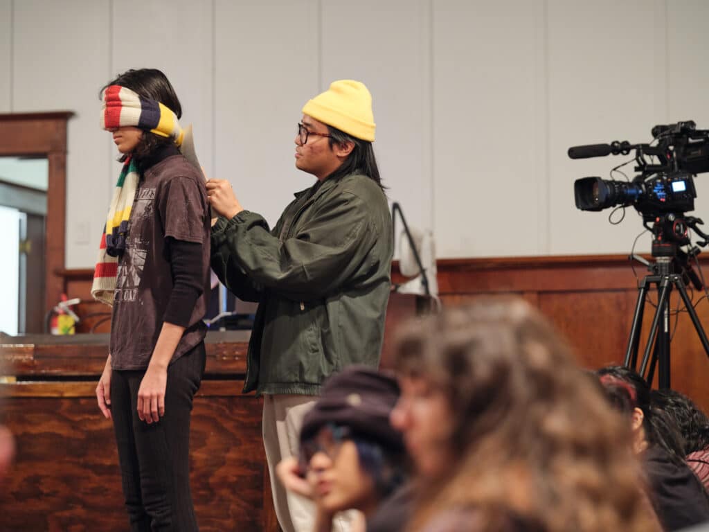 One person stands still with their eyes covered by a striped scarf while another gently adjusts the fabric behind their head. The quiet action unfolds in a wood-panelled room as seated audience members watch nearby.