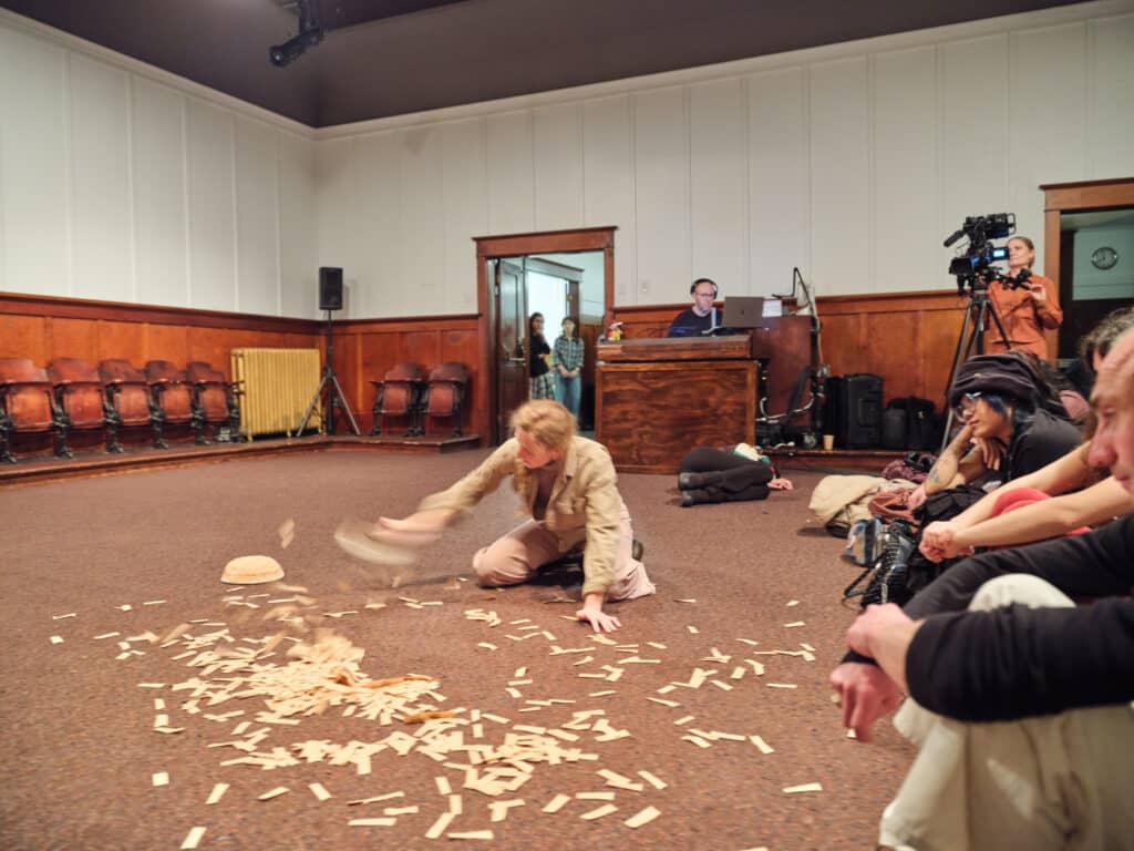 At the centre of a large room, a performer kneels on the floor scattering thin wooden fragments in a sweeping motion. Audience members sit close by in a wide circle, observing the unfolding performance.