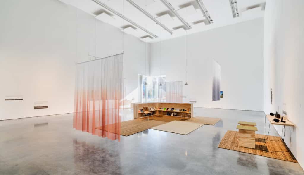 A bright, open gallery holds soft hanging curtains, woven rugs, and low wooden seating arranged around books. Light spills across the floor, shaping a quiet space for reading, listening, and lingering.
