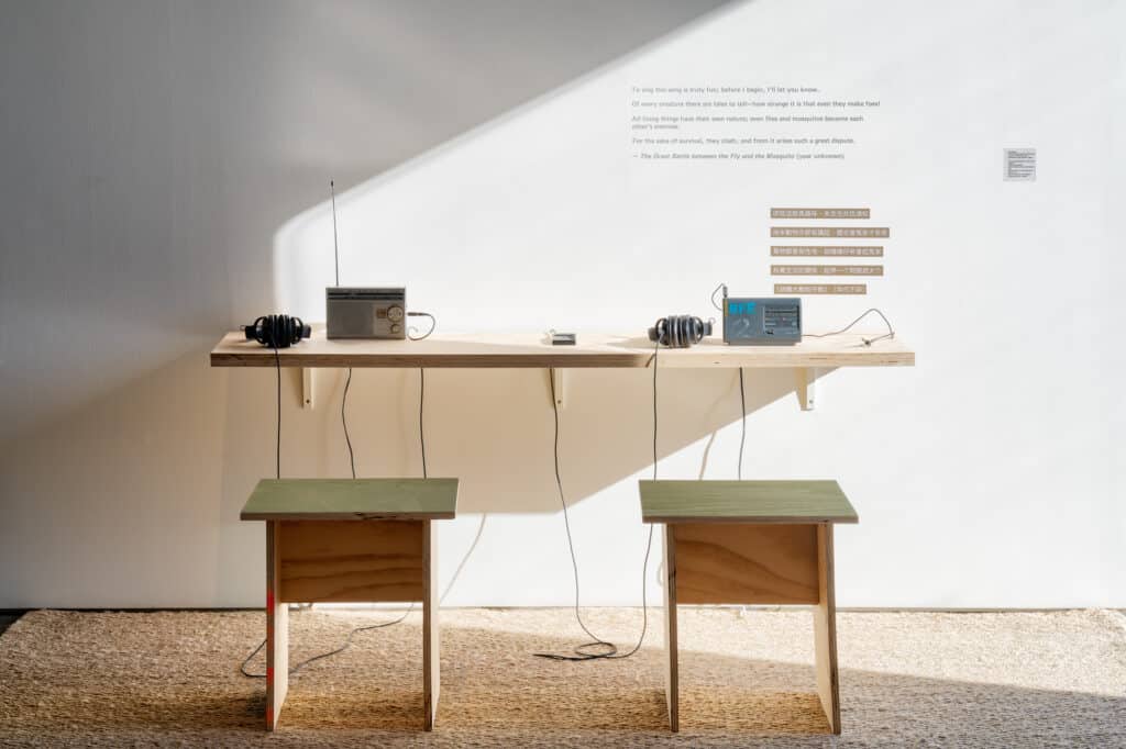 Two small stools face a simple wooden shelf with radios and headphones, their cords trailing downward. Sunlight cuts across the wall, illuminating text and inviting a moment of listening in stillness.