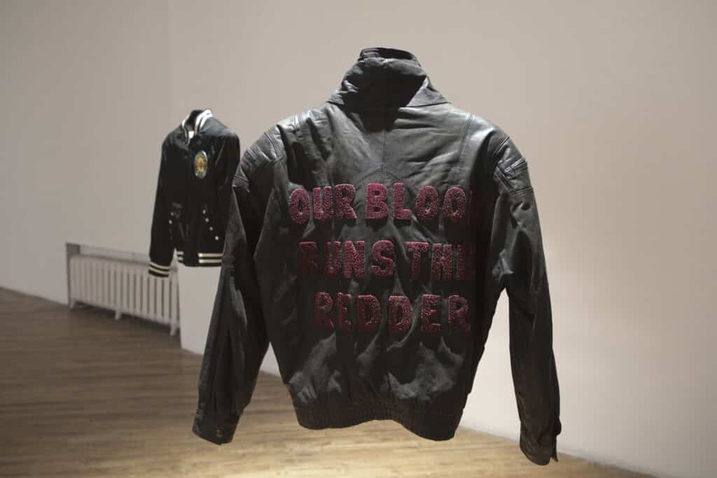 In a gallery space, a black leather jacket appears suspended in midair, its back embroidered with red beaded words reading “Our blood runs the redder.” Another jacket hangs softly in the background against a white wall.