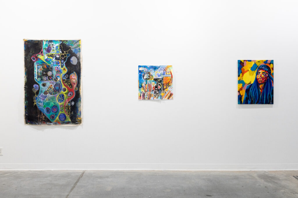 Three paintings hang on a gallery wall: a large colourful abstract on the left, a smaller mixed-media piece in the centre, and a bold portrait on the right